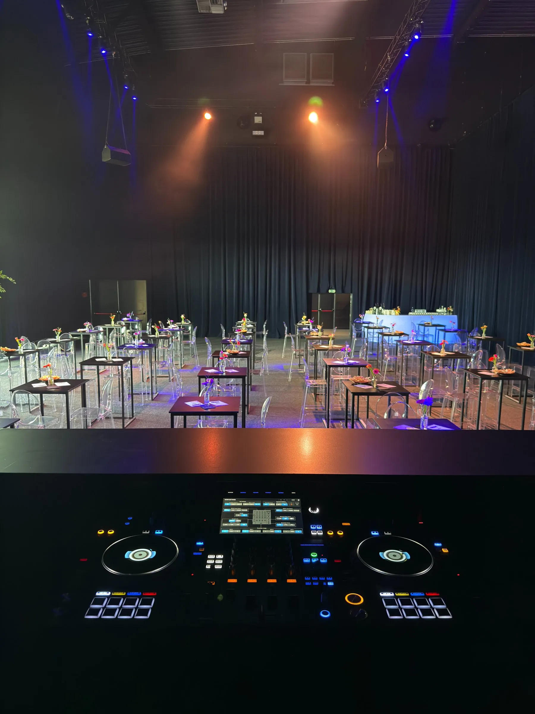 Zicht vanachter de DJ-booth op een zaal met statafels en spots