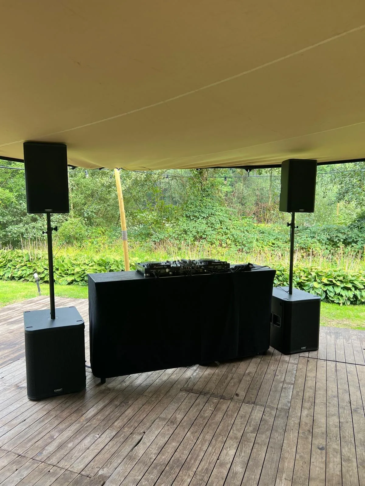 DJ-booth op een houten terras onder een feesttent, met zicht op de tuin